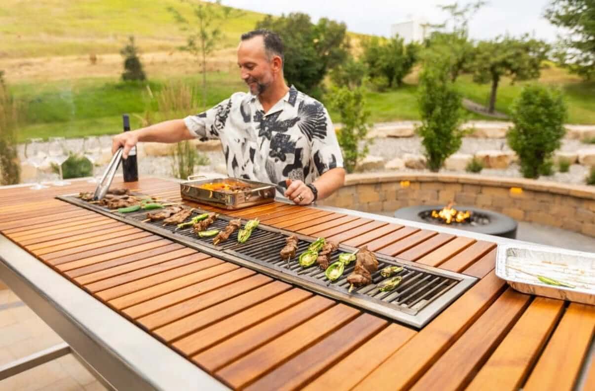  A man in a floral shirt is smiling while grilling meat and vegetables on an Angara BBQ table. He is using tongs to turn food on the long, built-in grill that is integrated into the center of the wooden table.