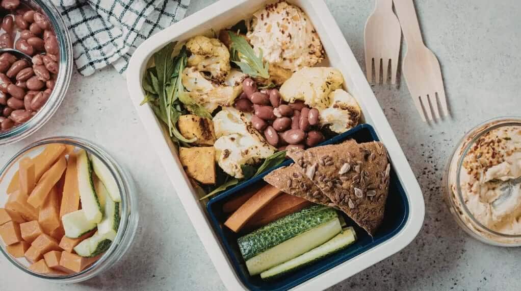 Plant-based lunch box with roasted cauliflower, kidney beans, arugula, hummus, cucumber, carrot sticks, and seeded crispbread for healthy meal delivery.