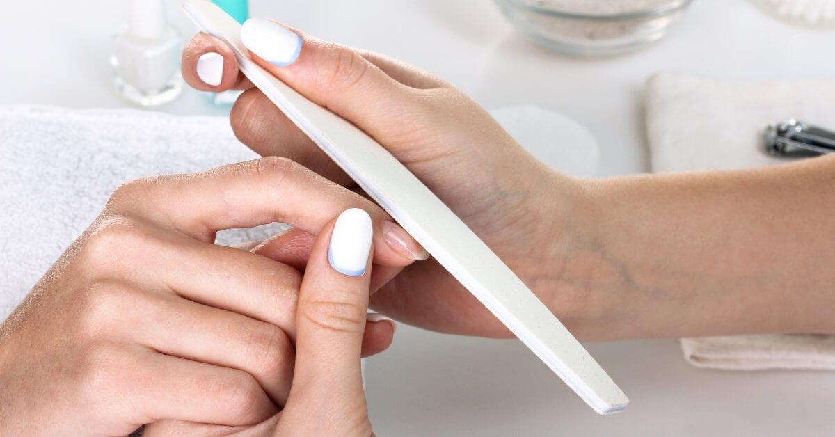 A close-up of a professional nail tech filing a customer's nail bed. The nail tech's nails are white and blue.