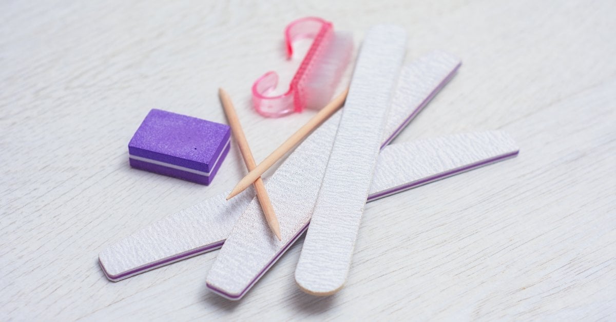 A close-up of a manicure kit featuring nail files, buffers, sticks, and a brush on a light-gray wooden background.