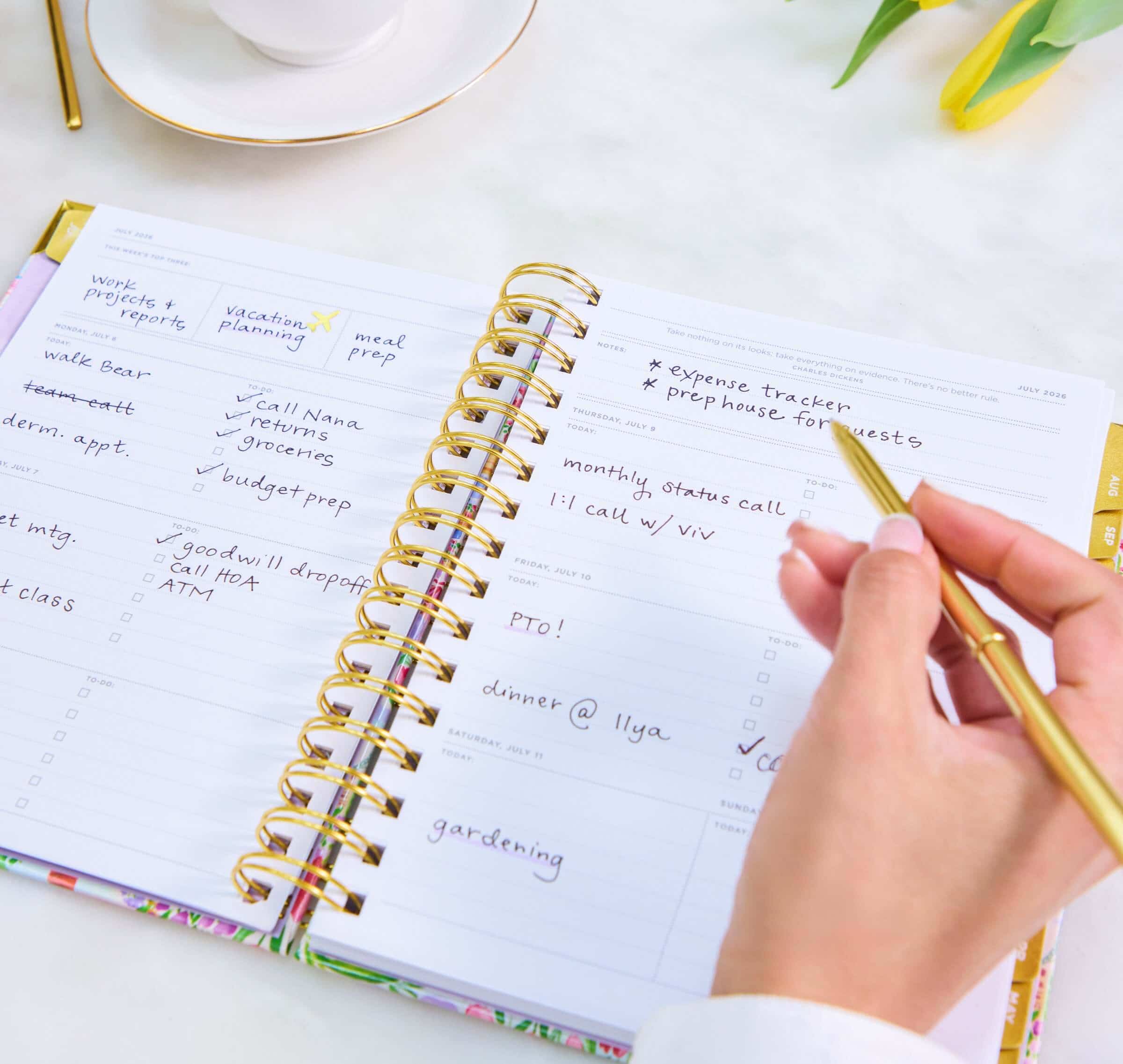 planning your academic year, hand holding gold pen, open book planner showing written on pages, coffee cup, yellow flowers, white background
